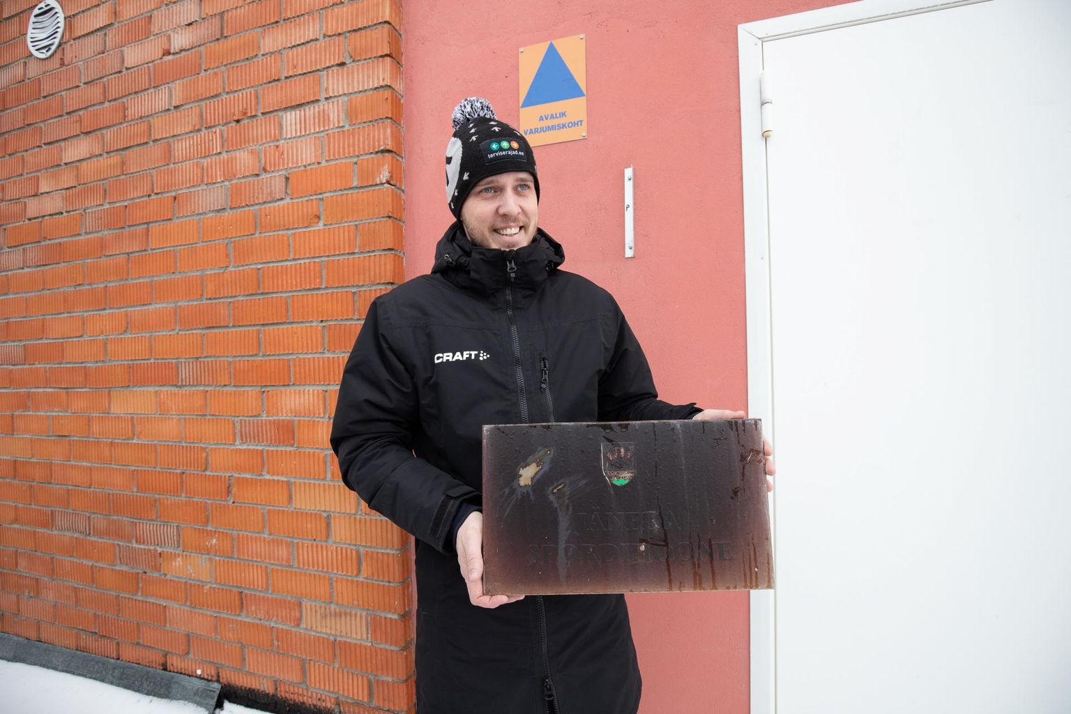 Spordihoone mustaks pleekinud silt Priit Hundi käes. Märk tema taga seinal annab teada, et hoone on ühtlasi avalik varjumiskoht.