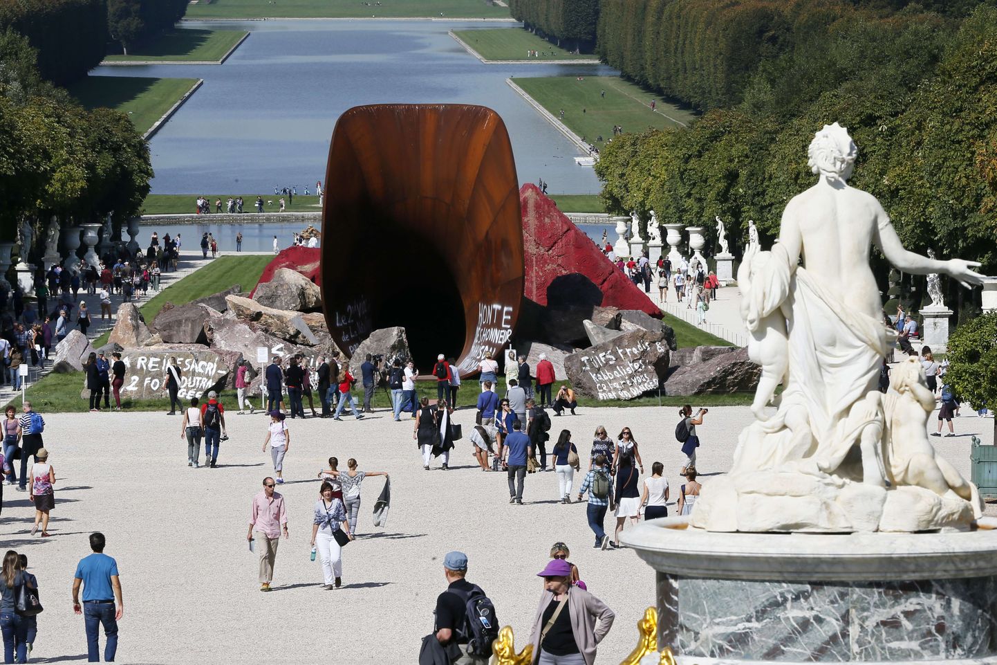 Visitors walk past the earth and mixed media monumental artwork "Dirty Corner" a 2011 Cor-Ten steel, by British contemporary artist of Indian origin Anish Kapoor on September 11, 2015 in Versailles at the Chateau of Versailles.  On September 6 anti-Semitic inscriptions and phrases such as "Queen sacrificed, twice insulted," "the second rape of the nation by deviant Jewish activism" and "Christ is king in Versailles" were sprayed in white paint on the sculpture. On September 1, the sculpture was vandalized for a third time, with graffiti saying "Respect Art as U trust God" scrawled on it, prompting he wanted the graffiti to remain to bear witness to hatred, thus opening a debate which could fall fowl of French laws repressing anti-Semitism. AFP PHOTO / PATRICK KOVARIK
