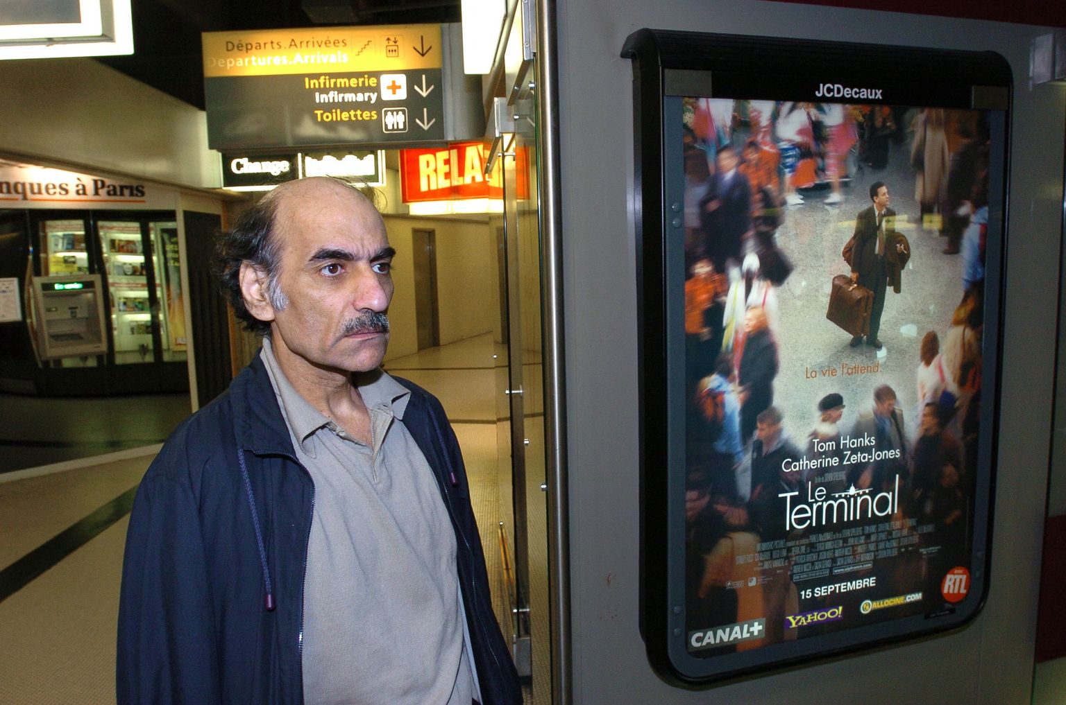 Mehran Karimi Nasseri 2004 Pariisi Charles De Gaulle'i lennujaamas, kus ta elas aastaid. Pildi on ta filmi «Terminal», mille inspiratsiooniks ta oli,  plakati juures