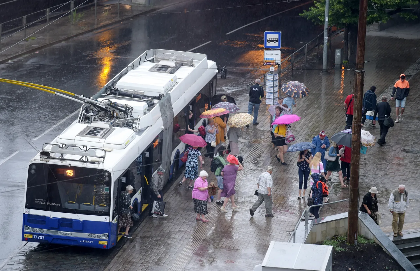 Cilvēki sabiedriskā transporta pieturvietā "Centrālā stacija" lietus laikā.
