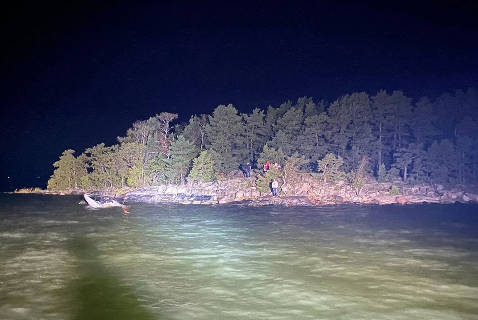 Päästeoperatsioon Naantali lähedal. Saare ranna lähedal vees on näha uppunud mootorpaadi targakaar, kaldal ootavad saarele pääsenud inimesed.