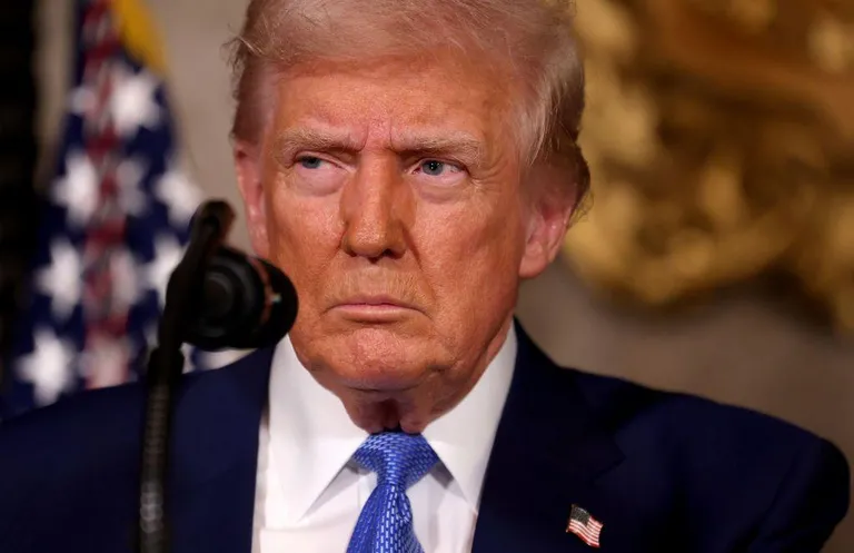 Трамп стоит на подиуме для микрофона, одетый в темно-синий костюм, белую рубашку и синий галстук.