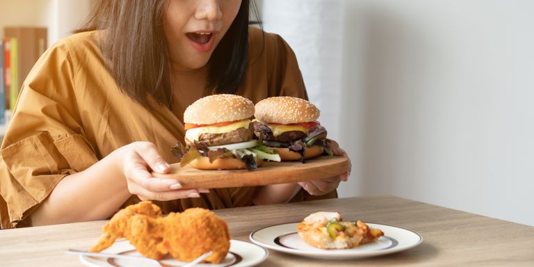 Голодная полная женщина, держащая гамбургеры на деревянной тарелке, и готовая съесть все остальное на столе. Концепция расстройства пищевого поведения.