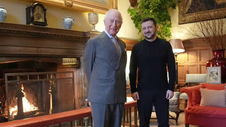 Король Карл III в сером костюме стоит у камина с Владимиром Зеленским в черной военной форме.