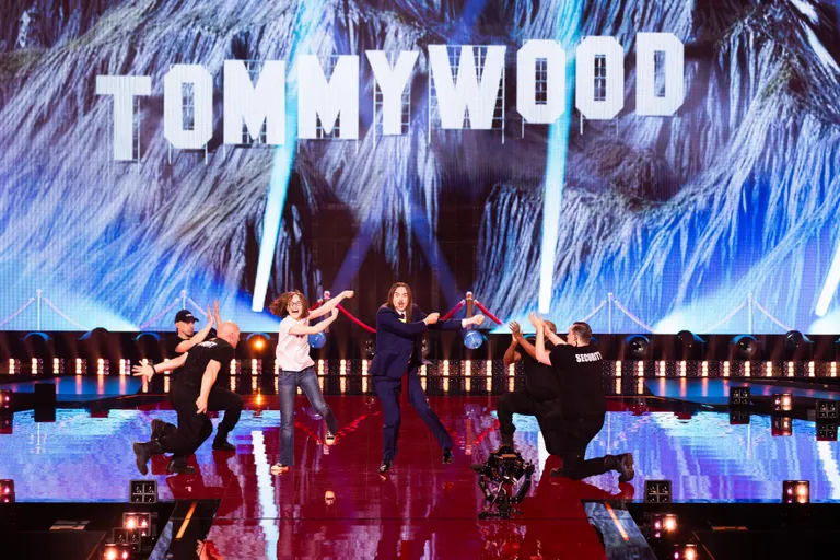 Tommy Cash vihtumas Eurovisiooni lauluvõistluse poolfinaalis tantsu andunud fänni mängiva naisega, artisti turvamehed aplodeerimas.