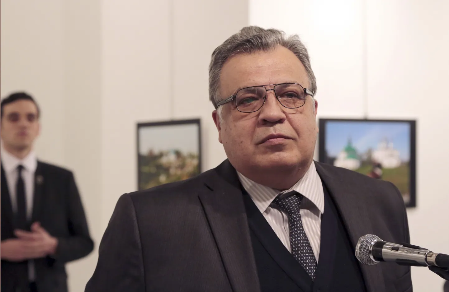 Andrei Karlov, the Russian Ambassador to Turkey, speaks at a photo exhibition in Ankara on Monday, Dec. 19, 2016, moments before a gunman opened fire on him. Karlov was rushed to a hospital after the attack and later died from his gunshot wounds.  The gunman is seen at rear on the left.  (AP Photo/Burhan Ozbilici)