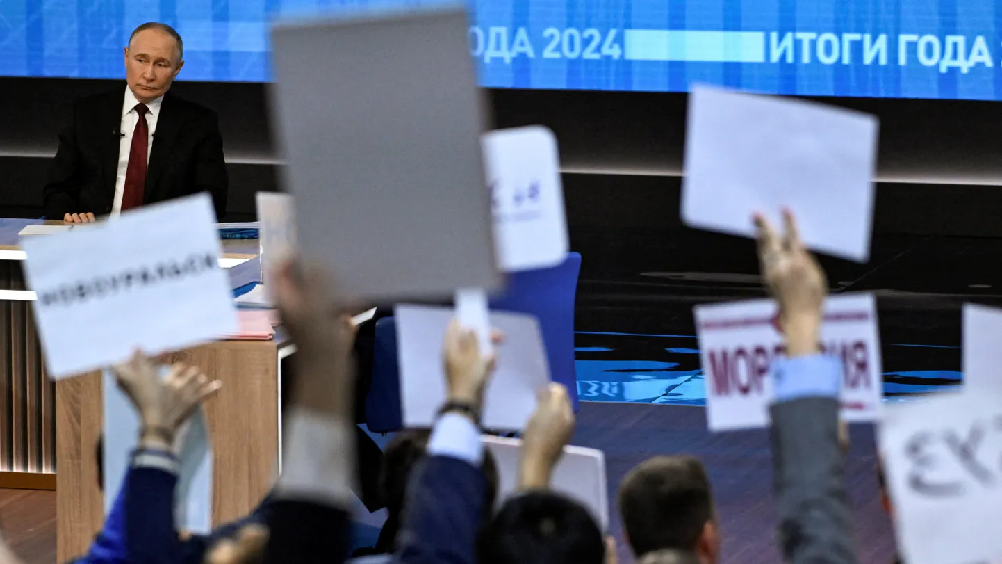 Пути сидит на фоне экрана с надписью «Итоги года 2024», перед ним - поднятые руки журналистов с листками в формате A4.