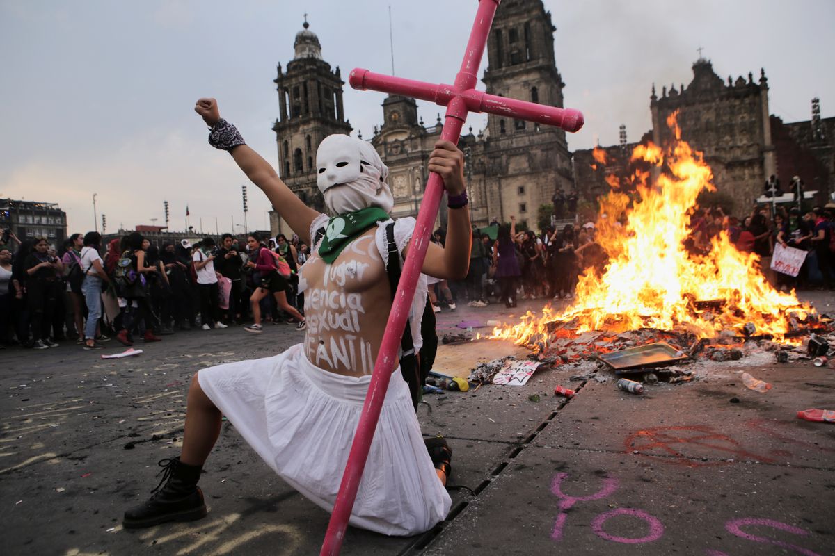 2020. gada 8. marts, Mehiko, Meksika. Protestā pret vardarbību pret sievietēm sieviete tur rokās rozā krustu. Aiz viņas deg ugunskurs, bet vārdi uz viņas kailā ķermeņa nozīmē - 
"Meksika ir seksuālās vardarbības valstu pirmajā vietā".