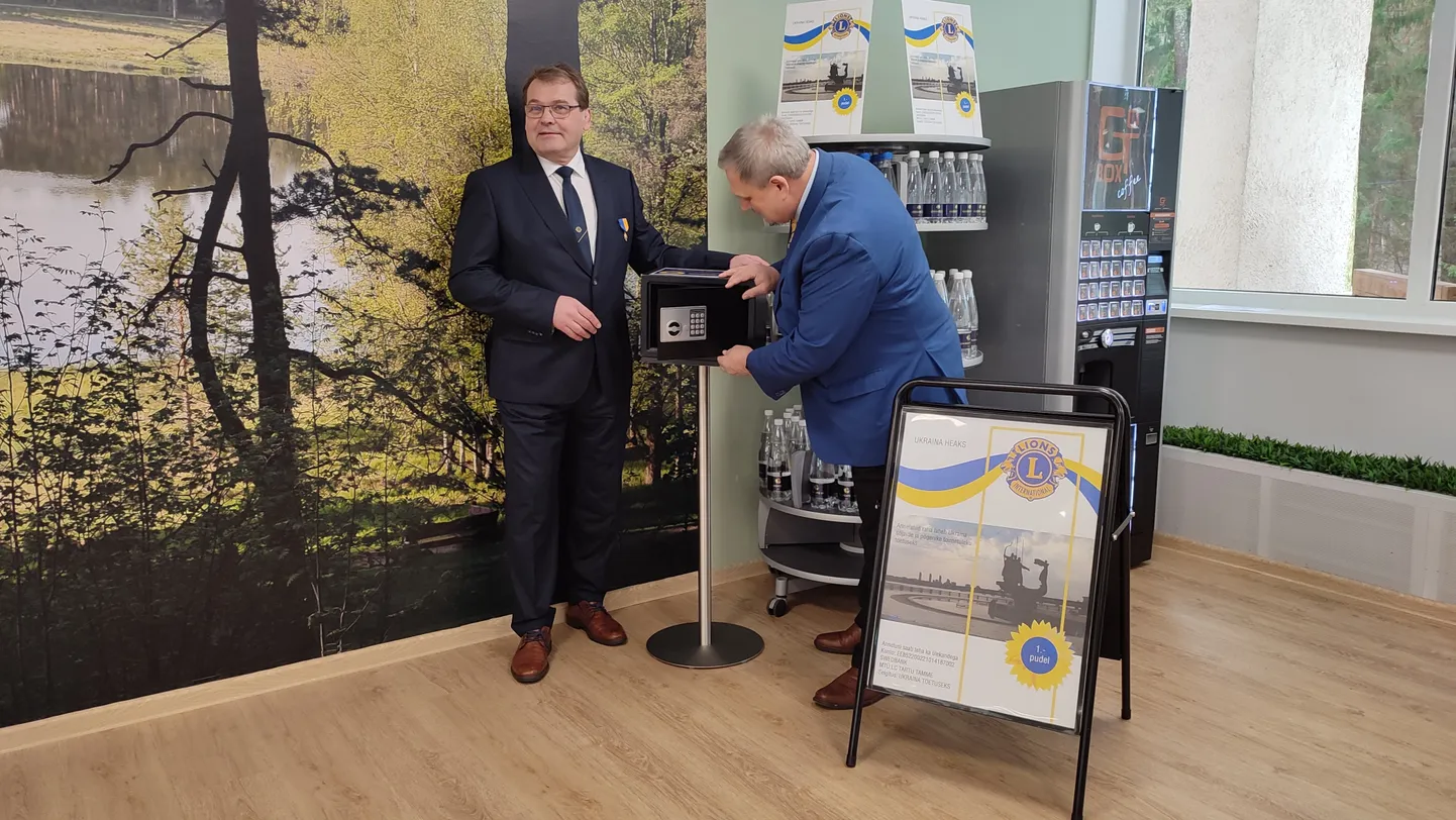 Elva haigla juhataja Peeter Laasik (vasakul) ja Tartu Tamme Lions-klubi president Andrus Kõre korrastavad haigla registratuuri ootesaalis asuvat Kuningate Vee hoidikut, mille abil kogutakse annetusi Ukraina toetuseks.